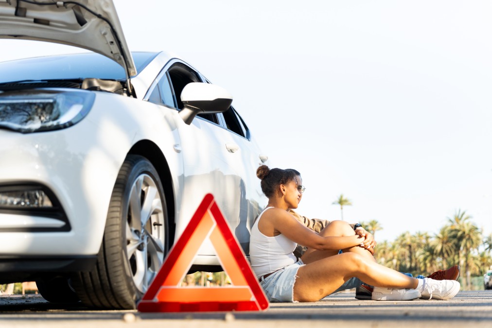Jeune femme assise près de sa voiture blanche en panne avec un triangle de signalisation, illustrant la nécessité de financer un imprévu sereinement.